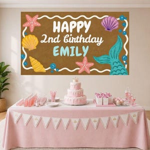 May include: A birthday party scene with a pink tablecloth, a banner, and a three-tiered cake. A large brown banner displays the text "HAPPY 2nd birthday EMILY" with mermaid and seashell illustrations. Balloons and party supplies are also visible.