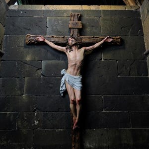 Può includere: Un crocifisso in legno con una figura dettagliata di Gesù con corona di spine, che indossa un perizoma grigio chiaro. La croce ha un aspetto strutturato simile alla corteccia con le lettere INRI incise in alto, montata su una parete di pietra scura.