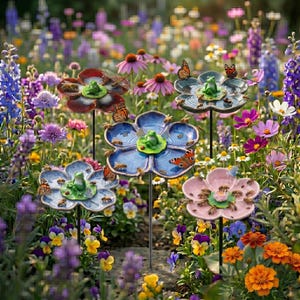 May include: Four ceramic flower-shaped garden stakes, each featuring a small green frog figurine at the center. The flowers are colored in deep red, blue, light blue, and pink, mounted on thin metal rods amidst a vibrant garden of various flowers.