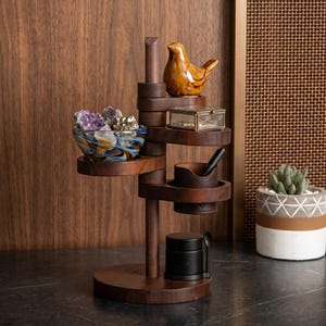 May include: A multi-tiered wooden organizer with various items. It features a small ceramic bird, a glass box, a leather pen holder, a black jar, and a decorative bowl filled with crystals. The organizer is on a dark surface.