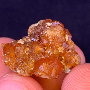 Op de afbeelding: Close-up van een cluster van oranje-bruine kristallen. De kristallen hebben een geometrische vorm en zijn geclusterd. Het mineraal heeft een ruwe textuur en wordt vastgehouden door de vingers van een persoon. De achtergrond is donker.