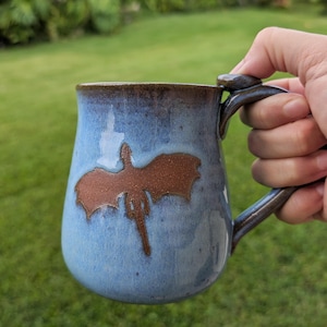 May include: A ceramic mug with a light blue glazed finish and a dark brown rim. The mug features a textured, dark brown silhouette of a dragon with outstretched wings on the front. It has a sturdy, dark brown handle attached to the side.