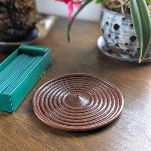 May include: A brown ceramic incense holder with a concentric circle design. A green box of incense sticks is visible to the left. The incense holder is on a wooden surface. The image is well-lit and shows the details of the incense holder.