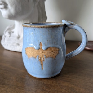 May include: A light blue ceramic mug with a speckled finish and a brown dragon silhouette embossed on the front. The mug features a curved handle with a small circular thumb rest at the top and a brown rim that matches the dragon design.
