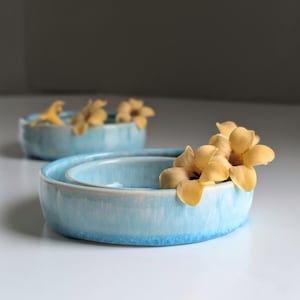 May include: Two light blue ceramic bowls, one nested inside the other, each holding yellow flowers. The bowls have a glossy finish and a slightly uneven rim. The background is a soft, neutral color.