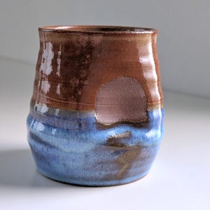 May include: A handmade ceramic tumbler with a two-tone glaze. The top half is a rich brown, while the bottom half is a vibrant blue. The cup has a slightly textured surface and a unique, organic shape. A perfect addition to any kitchen.