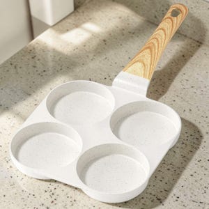 May include: White speckled egg frying pan with four circular molds. The pan has a light wood grain handle with a hole for hanging. The pan is designed for cooking multiple eggs or pancakes at once.
