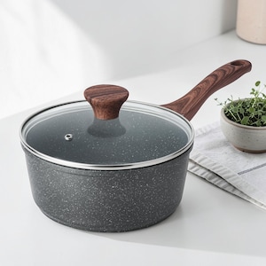 May include: A dark gray speckled saucepan with a clear glass lid and a wooden handle and knob. The handle and knob are made of a dark wood. The pan is sitting on a white surface.