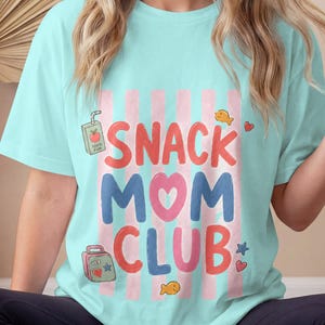 Könnte beinhalten: Ein hellblaues T-Shirt mit einem Grafikdruck mit vertikalen rosa Streifen und dem Text "SNACK MOM CLUB" in roten und blauen Buchstaben. Das Design enthält kleine Illustrationen einer Saftpackung, einer Brotdose, Goldfisch-Crackern und Herzen.