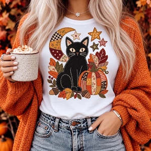 Op de afbeelding: Een wit t-shirt met een grafische print van een zwarte kat die naast een patchwork pompoen, een maansikkel en sterren zit. Het ontwerp bevat herfstachtige elementen zoals kleurrijke bladeren en eikels in tinten oranje, bruin en goud.