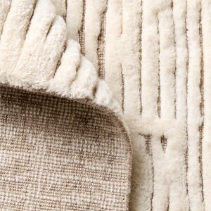 Puede incluir: Primer plano de una alfombra color crema con un patr&oacute;n de rayas verticales texturizadas. Se ve la parte inferior de la alfombra, que revela un reverso tejido marr&oacute;n. Los bordes de la alfombra est&aacute;n ligeramente curvados, mostrando el contraste.