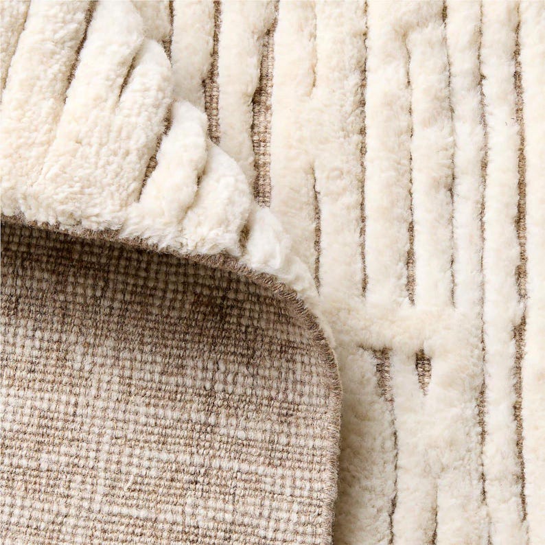 Puede incluir: Primer plano de una alfombra color crema con un patr&oacute;n de rayas verticales texturizadas. Se ve la parte inferior de la alfombra, que revela un reverso tejido marr&oacute;n. Los bordes de la alfombra est&aacute;n ligeramente curvados, mostrando el contraste.