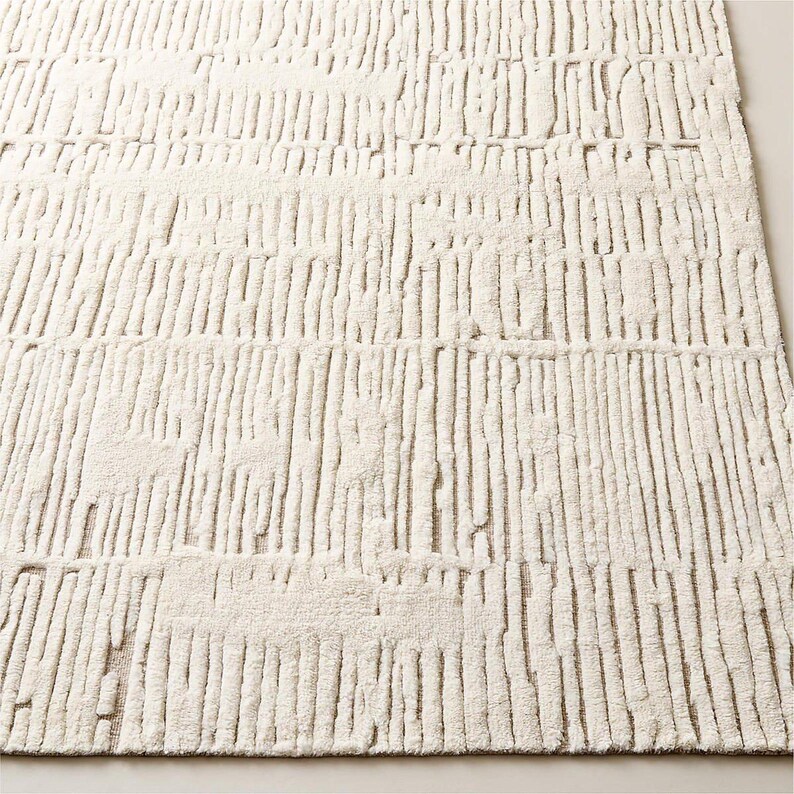 Puede incluir: Alfombra rectangular de color crema con una superficie texturizada. La alfombra presenta un patr&oacute;n de l&iacute;neas verticales, creando un efecto rayado. Las l&iacute;neas est&aacute;n ligeramente elevadas, agregando profundidad e inter&eacute;s visual al dise&ntilde;o de la alfombra. La alfombra est&aacute; hecha de un material suave y afelpado.