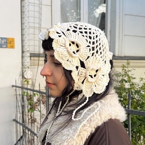 May include: Cream-colored crochet hat with large floral accents. The hat features a delicate open weave design and is adorned with three-dimensional flowers. The hat is worn with a brown jacket with a cream-colored, fluffy collar.