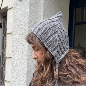 May include: A grey knitted hat with a pointed crown and braided ties. The hat features a ribbed knit texture and a chevron pattern on the front. This neutral-toned hat is suitable for autumn and winter wear.