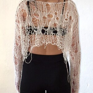 Peut inclure: Haut en maille ajour&eacute;e blanc cass&eacute; &agrave; manches longues. Le haut pr&eacute;sente un design d&eacute;chir&eacute; et effiloch&eacute; avec de grands trous. Le mod&egrave;le porte un soutien-gorge noir et un pantalon noir. Le haut est en mati&egrave;re l&eacute;g&egrave;re.