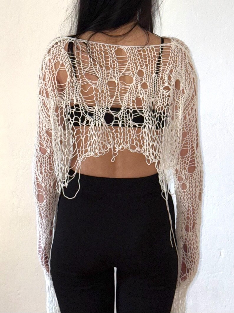 Peut inclure: Haut en maille ajour&eacute;e blanc cass&eacute; &agrave; manches longues. Le haut pr&eacute;sente un design d&eacute;chir&eacute; et effiloch&eacute; avec de grands trous. Le mod&egrave;le porte un soutien-gorge noir et un pantalon noir. Le haut est en mati&egrave;re l&eacute;g&egrave;re.