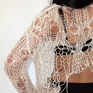 Peut inclure: Haut &agrave; manches longues en maille ajour&eacute;e blanc cass&eacute;. Le v&ecirc;tement pr&eacute;sente un motif en forme de filet avec de grands trous et un ourlet effiloch&eacute;. Le haut est port&eacute; sur un v&ecirc;tement noir, avec le dos et le bras visibles.