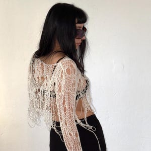 Peut inclure: Un haut en crochet cr&egrave;me &agrave; tissage ouvert, avec des manches longues et une coupe courte. Le haut pr&eacute;sente un design vieilli avec de multiples trous et des fils pendants. Le mod&egrave;le porte un pantalon noir et des lunettes de soleil.