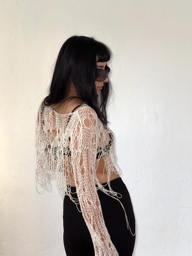 Peut inclure: Un haut en crochet cr&egrave;me &agrave; tissage ouvert, avec des manches longues et une coupe courte. Le haut pr&eacute;sente un design vieilli avec de multiples trous et des fils pendants. Le mod&egrave;le porte un pantalon noir et des lunettes de soleil.