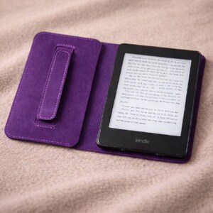 May include: A purple Kindle e-reader in an open, matching case. The e-reader displays text on its screen. The case has a hand strap and is made of a soft, suede-like material. The Kindle is on a neutral-colored surface.