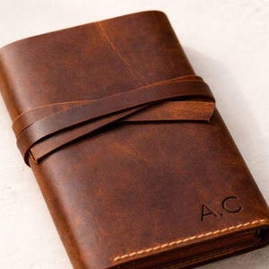 May include: A brown leather journal with a wrap-around strap closure. The journal has the initials "A.C." embossed on the front. The leather has a textured appearance and is stitched along the edges.