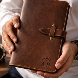 May include: A brown leather journal with a buckle closure and mountain graphic with the initials "A.B." embossed on the front. The journal is held by a person wearing a white shirt and a watch with a brown leather strap.