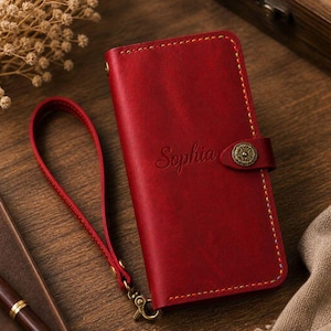 Puede incluir: Una funda roja de cuero para teléfono con el nombre "Sophia" grabado en relieve en la parte delantera. La funda tiene una correa de muñeca y un cierre decorativo. Las costuras son de color amarillo contrastante. La funda se cierra con un botón decorativo.