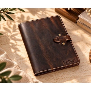 May include: A dark brown leather journal featuring a textured surface, light-coloured stitching along the edges, and a strap closure secured with two brass-coloured metal studs. The name Robert is embossed in the bottom right corner of the cover.