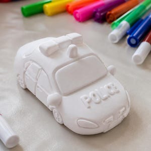 Könnte beinhalten: Eine weiße, unbemalte Polizeiauto-Figur aus Keramik mit dem eingeprägten Wort POLICE auf der Motorhaube, einem Dachlichtbalken und Seitenspiegeln, umgeben von einer Vielzahl bunter Filzstifte, die zum Ausmalen und Dekorieren bereitliegen.