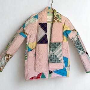 May include: A quilted jacket constructed from various fabric patches in shades of pink, blue, yellow, and green. The garment features a shawl collar, long sleeves, and a patchwork design with geometric and floral patterns. A label is visible at the neck.