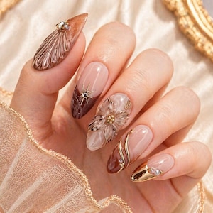 May include: A hand with long, almond-shaped nails, each adorned with intricate designs. The nails feature a mix of nude, brown, and gold tones, with embellishments like rhinestones, floral accents, and metallic swirls. The overall aesthetic is elegant and glamorous.