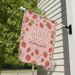 以下が含まれることがあります： ピンク色のガーデンフラッグ。イチゴのパターンと白い文字で「Sweet Summer strawberry garden」と書かれています。赤いイチゴ、緑の葉、小さな白い花で飾られています。白いポールに吊るされています。