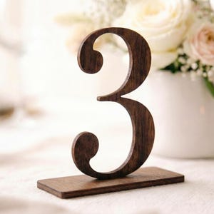 Può includere: Un numero da tavolo in legno marrone scuro, il numero tre, in piedi su una base rettangolare. Il numero è in un carattere stilizzato. Lo sfondo include un vaso bianco con fiori.