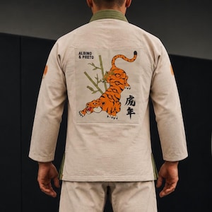 Puede incluir: Una chaqueta de gi para artes marciales de color beige con un gran parche cuadrado en la espalda. El parche muestra un tigre naranja trepando por tallos de bambú verdes, el texto "ALBINO & PRETO" en negro y kanjis japoneses a la derecha.