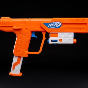 Op de afbeelding: Oranje en witte speelgoedpistool met het woord "NERF" in blauw. Het speelgoedpistool heeft een blauwe trekker en een wit magazijn. De grip heeft een textuurpatroon. Het speelgoedpistool staat tegen een zwarte achtergrond.