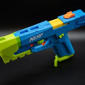 Może przedstawiać: Niebieski, żółty i zielony pistolet zabawkowy Nerf. Na pistolecie zabawkowym wydrukowano napis "NERF". Pistolet zabawkowy ma pomarańczowy przycisk na górze i zieloną końcówkę.