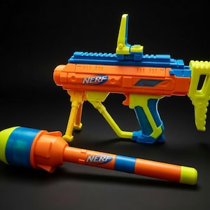 Op de afbeelding: Een oranje, blauw en geel Nerf speelgoedgeweer en projectiel. Het geweer heeft een vizier, een tweepoot en een handvat. Het projectiel is oranje met blauwe en gele strepen. Het Nerf-logo is zichtbaar.