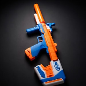 May include: A toy blaster gun in orange, blue, and white. The toy gun has a long orange barrel, a blue handle, and a white and blue stock. The word "NERF" is printed on the stock.