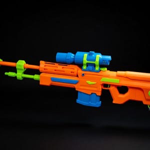 Op de afbeelding: Een felgekleurd speelgoedgeweer in oranje, blauw en groen. Het speelgoedgeweer heeft een vizier en andere realistische details. Het speelgoed is ontworpen om op een sluipschuttersgeweer te lijken.