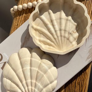 May include: Two off-white, seashell-shaped wooden bowls. One is open, the other a lid or decorative piece. They sit on a light grey surface. A wooden beaded garland is in the background. The bowls are likely for decorative use.