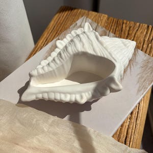May include: A white ceramic seashell-shaped decorative bowl. The bowl has a textured surface and a large opening. It sits on a white surface with a wooden background and a beige fabric in the foreground. The seashell is a decorative item.
