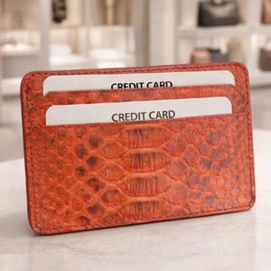 Op de afbeelding: Een oranje leren kaarthouder met een reptielhuidtextuur. Twee creditcards zijn zichtbaar in de sleuven, elk met de tekst "CREDIT CARD" erop. De kaarthouder is rechthoekig en heeft afgeronde hoeken.