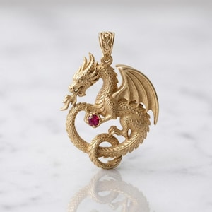 Könnte beinhalten: Ein goldener Anhänger in Form eines Drachen mit detaillierter Schuppenstruktur und texturierten Flügeln. Der Drache ist kreisförmig gebogen und hält einen kleinen, runden, leuchtend roten Edelstein in seiner Vorderkralle. Oben befindet sich eine Öse.