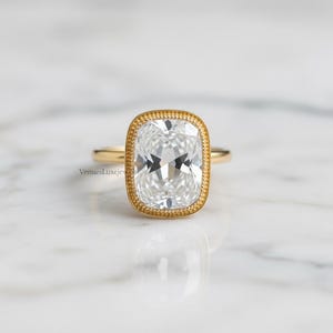 May include: A gold ring featuring a large, rectangular, clear gemstone with a halo setting. The ring's band is thin and gold, and the gemstone is set in a gold bezel. The ring is displayed on a white surface.
