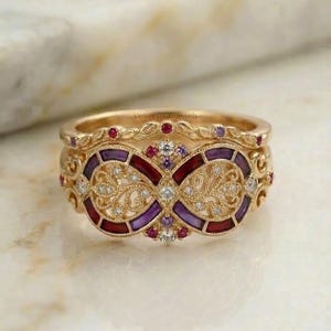 May include: A set of gold-toned rings with intricate filigree and gemstone details. The main ring features a central infinity symbol design with red and purple enamel, surrounded by small diamonds and gemstones. The top ring has a delicate leaf pattern.