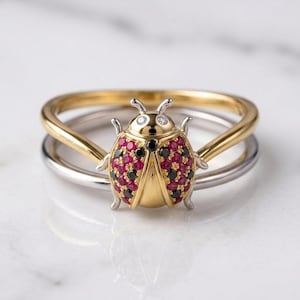May include: A two-tone ring featuring a gold and silver band with a ladybug centerpiece. The ladybug has a gold head with two small clear stones, and wings encrusted with red and black stones. The text VenuesLuxeJewels is visible on the band.