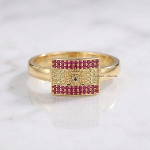 May include: A gold ring featuring a rectangular face set with red and yellow stones arranged in a horizontal striped pattern resembling the Spanish flag, with a central crest design and the text VenuesLuxeJewels visible on the side.