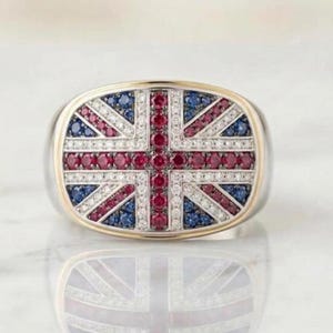 May include: A silver-toned ring featuring a rectangular face inlaid with a Union Jack flag pattern. The design is composed of small, round, pavé-set red, white, and blue gemstones arranged in the iconic cross and diagonal stripes of the British flag.
