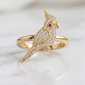 May include: A gold ring featuring a cockatoo bird motif encrusted with small white pavé diamonds and a single round red gemstone for the eye. The band is smooth and polished gold. The text VenuesLuxeJewels is visible on the ring band.